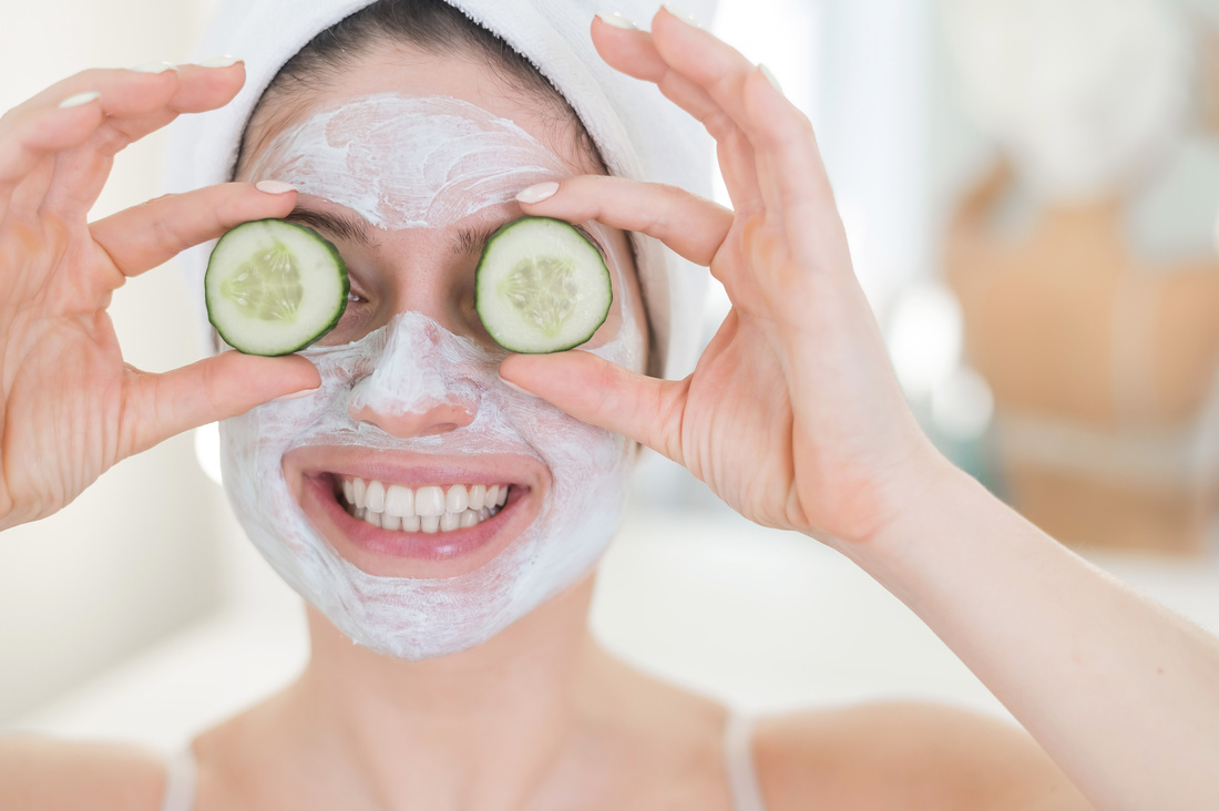 Cheerful Woman with a Towel on Her Hair and in a Clay Face Mask and Cucumbers on Her Eyes. Taking Care of Beauty at Home